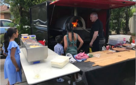 Joe cooking in the wood-fired oven on the trailer while children watch at a birthday party